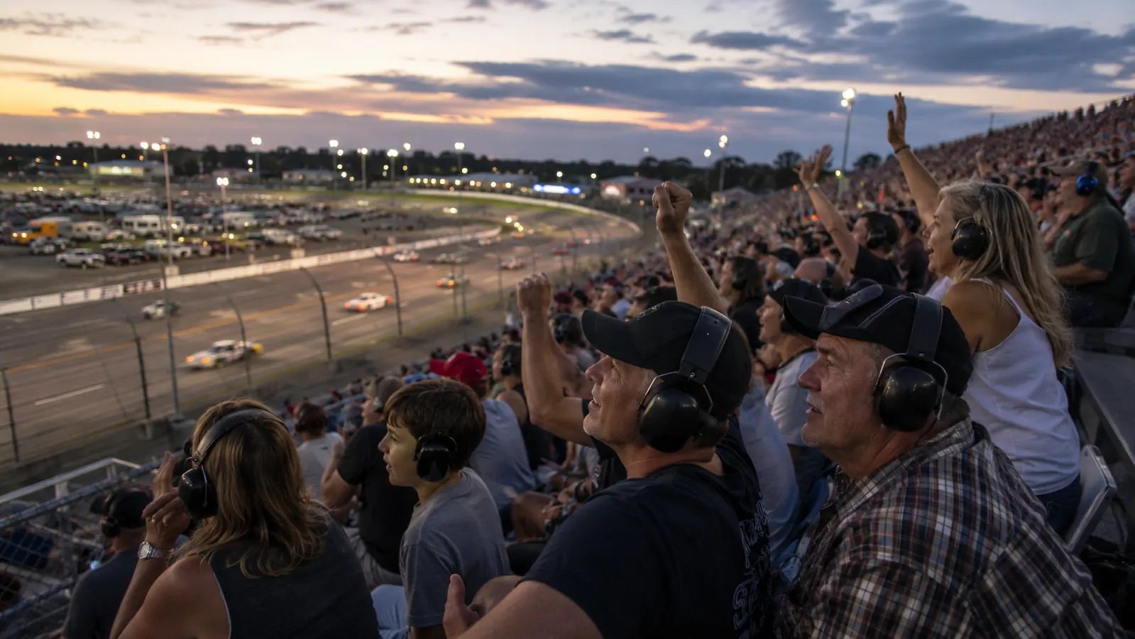 Crowd of race fans in grandstand seats cheering, several wearing black electronic earmuffs, cars racing below at dusk