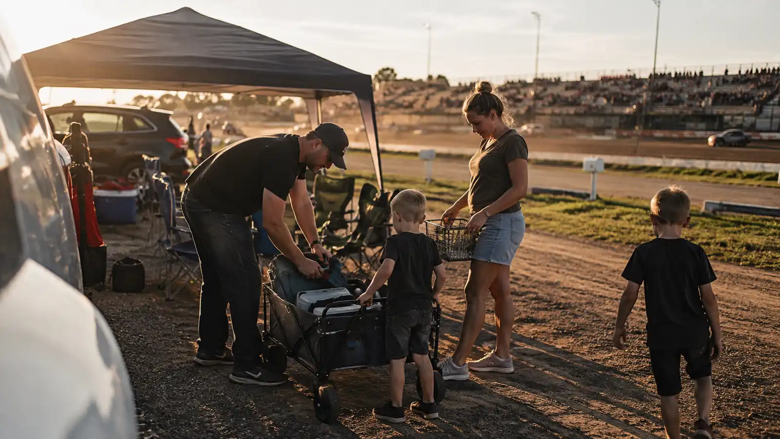 Family unloading a folding wagon at a dirt track tailgate spot with lawn chairs already set up