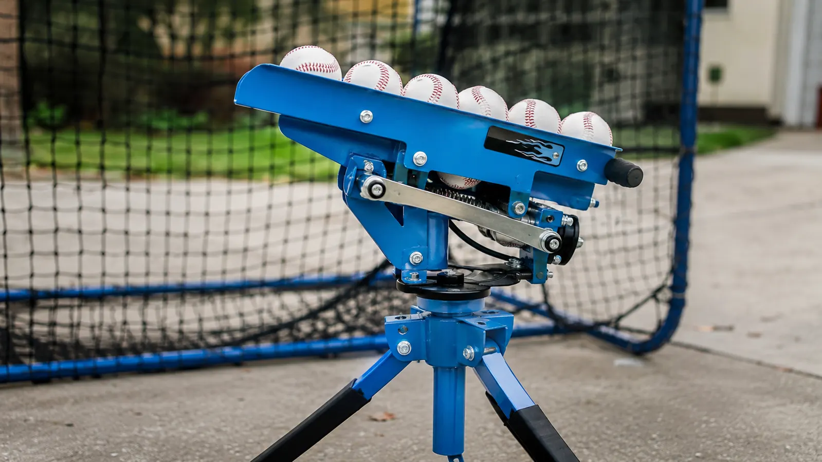 Close-up of a yellow pitching machine loaded with baseballs on a tripod in a backyard batting cage