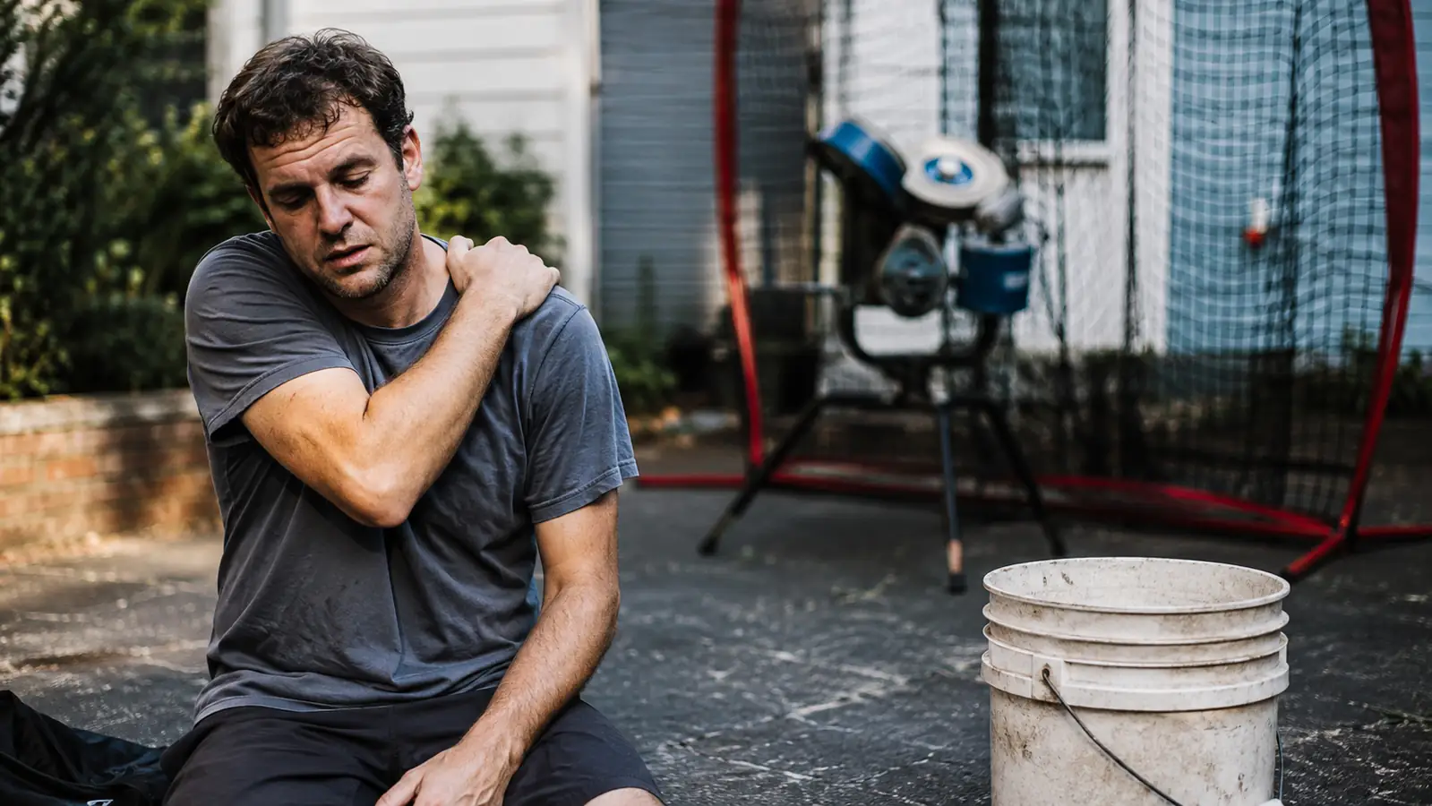 A man rubbing his shoulder while standing near a bucket of baseballs and a pitching machine in a driveway
