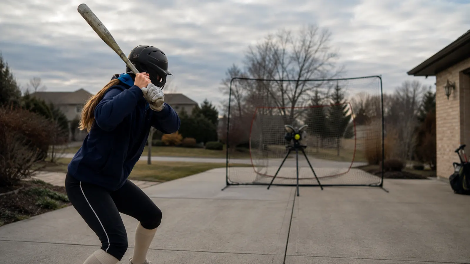Teenage softball player in batting stance facing a pitching machine set up in a driveway