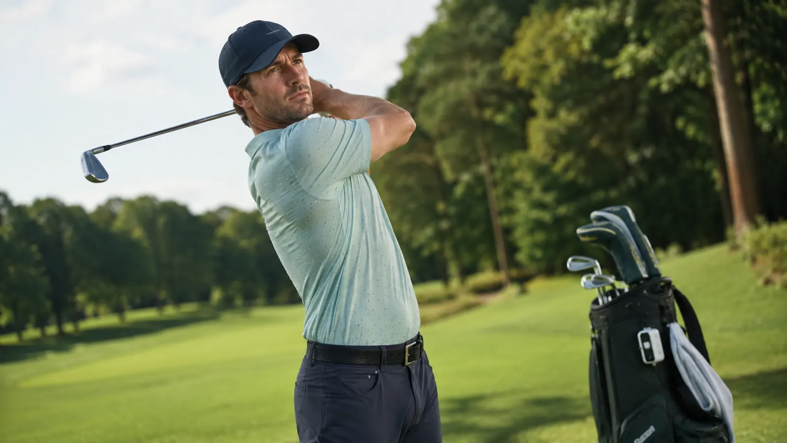 Golfer mid-swing on an approach shot toward a green with a rangefinder visible clipped to his bag in the background