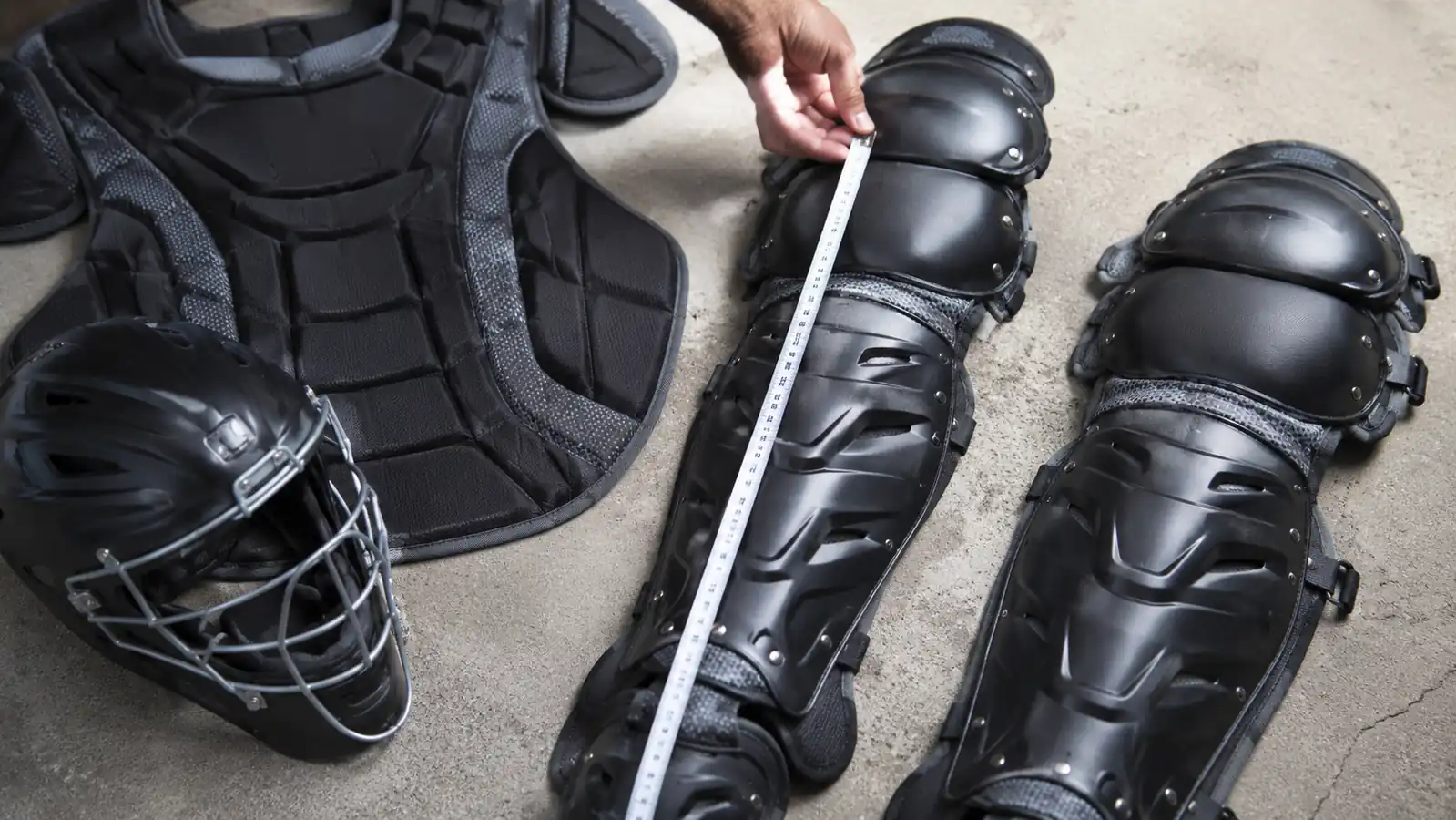 Hand holding a tape measure against a catcher's leg guard from the kneecap to the top of the ankle to check length