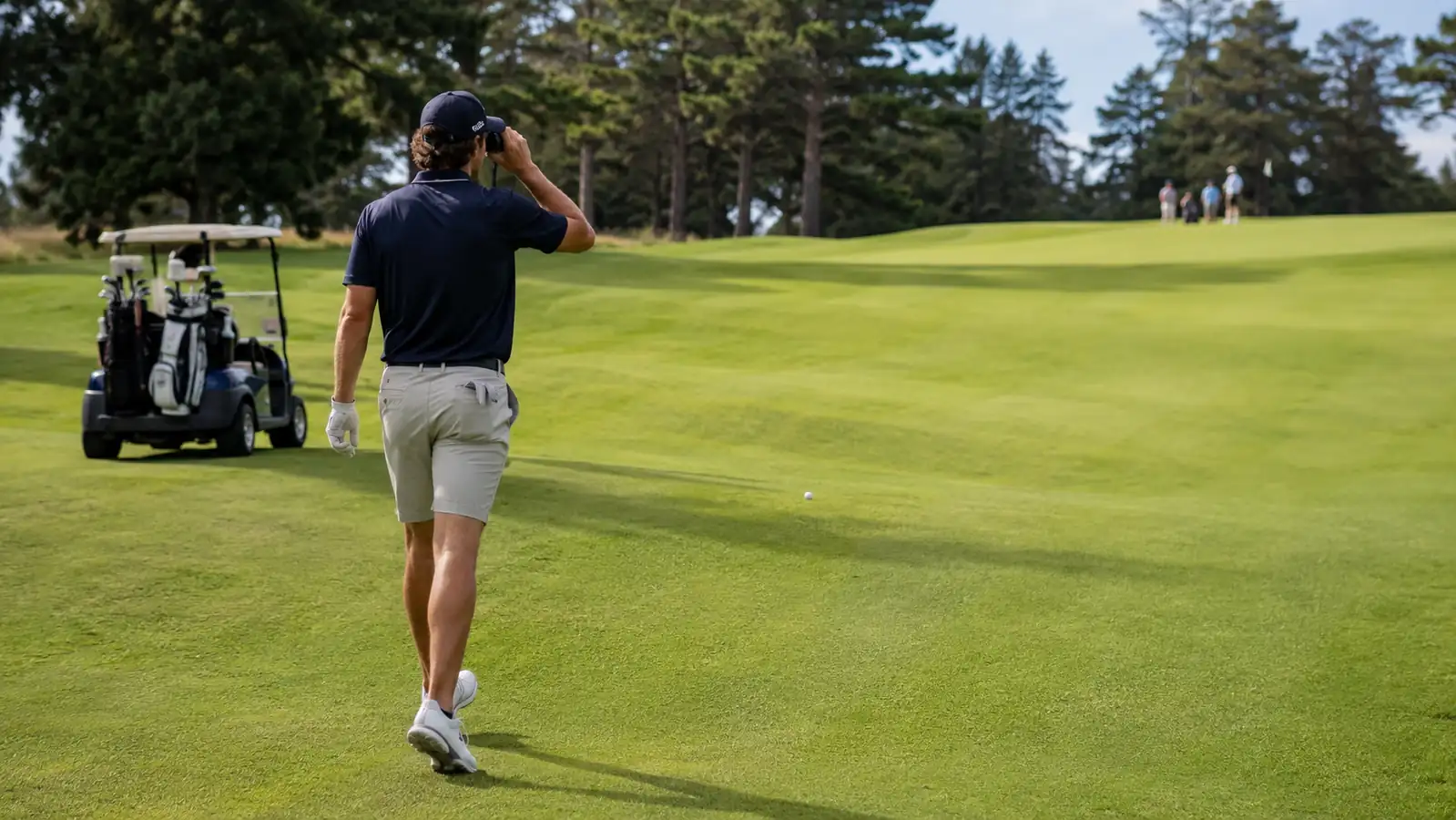 A golfer walking briskly up the fairway toward his ball while already raising the rangefinder to his eye