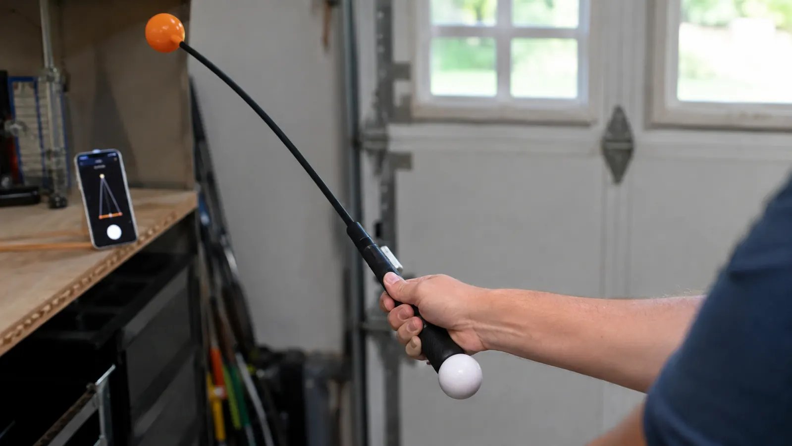 Hands gripping the swing trainer at address with a phone showing a metronome app propped on a nearby ledge