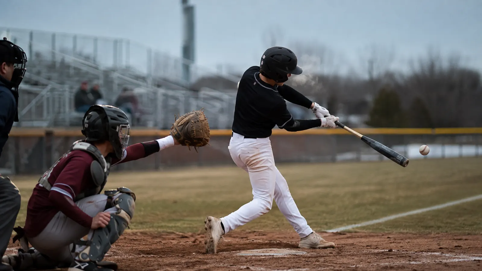 Teenage travel ball player mid-swing at an early spring tournament under cloudy skies