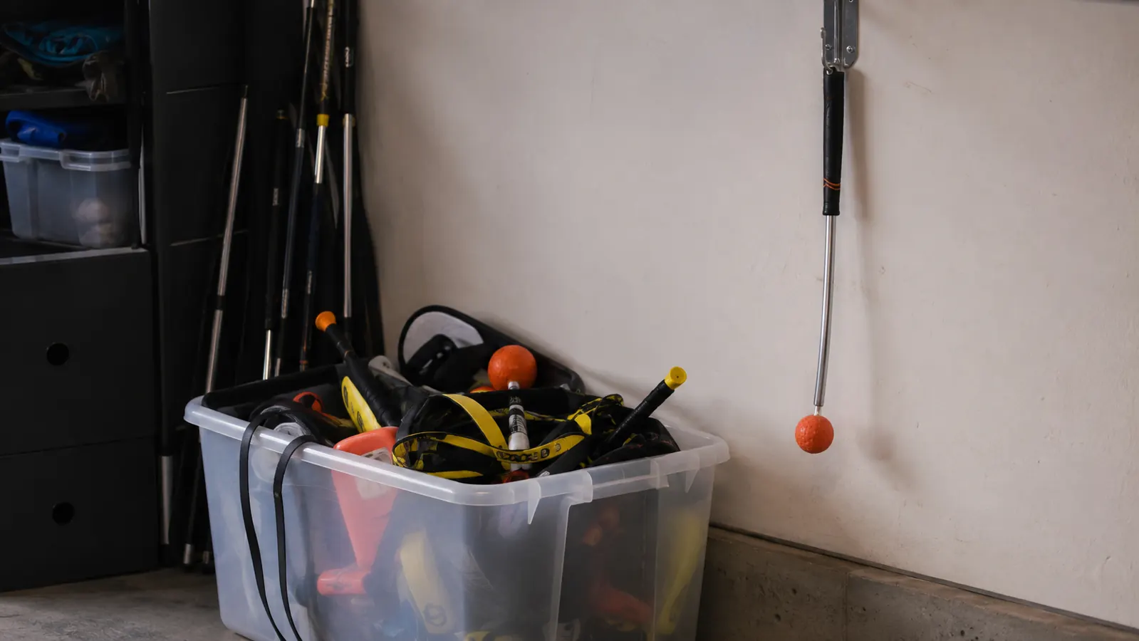 Storage bin of unused golf training gadgets sitting in a garage corner next to a wall-mounted training club
