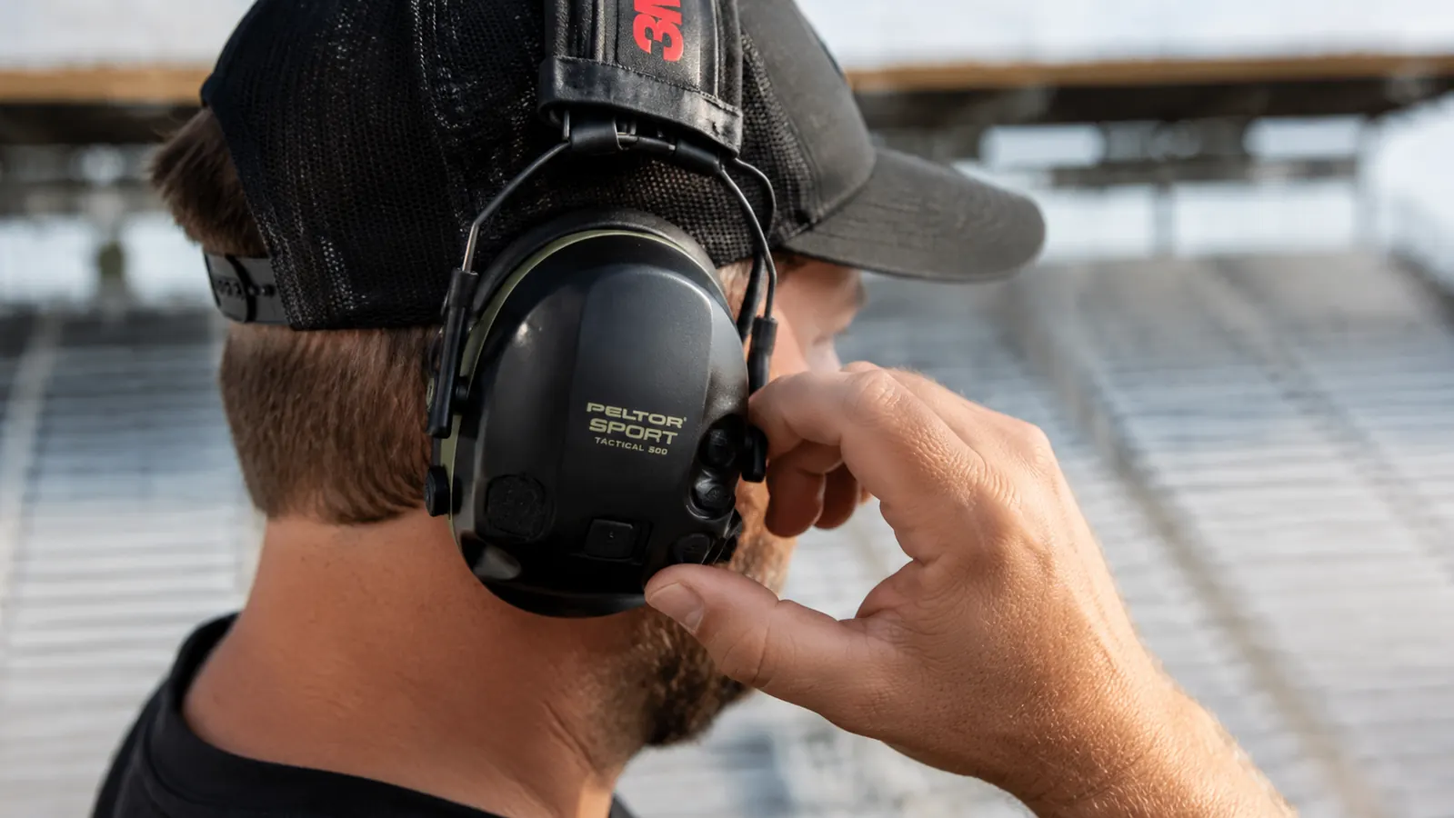 Hand adjusting the volume dial on a black electronic earmuff while wearing a ball cap at a race track