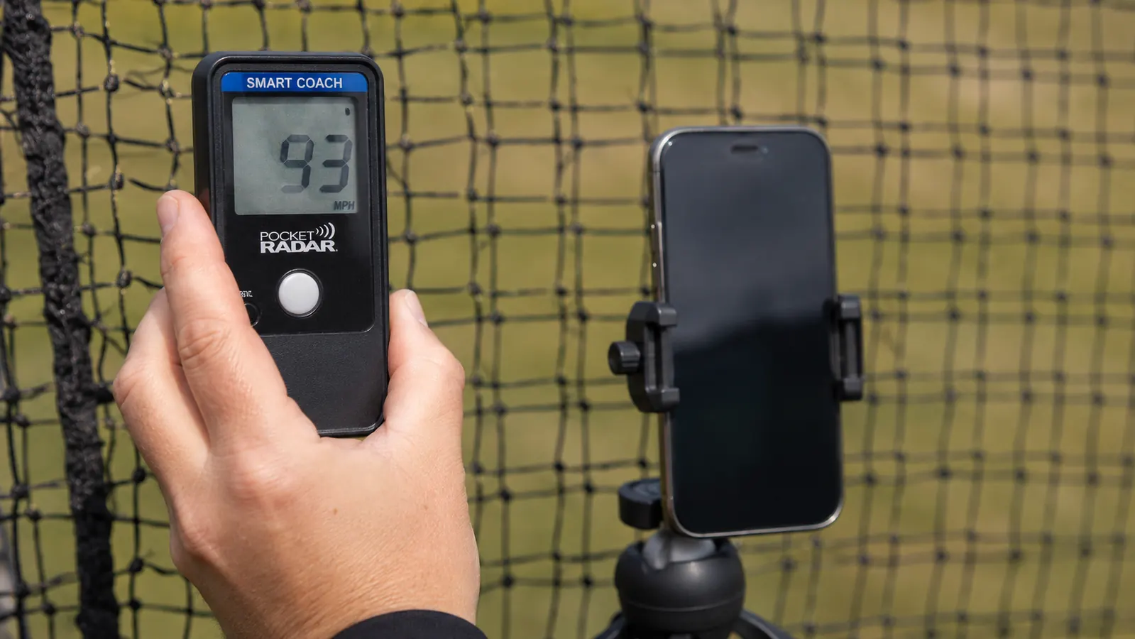 Close-up of a hand holding a Pocket Radar Smart Coach unit with a phone showing a velocity reading mounted nearby