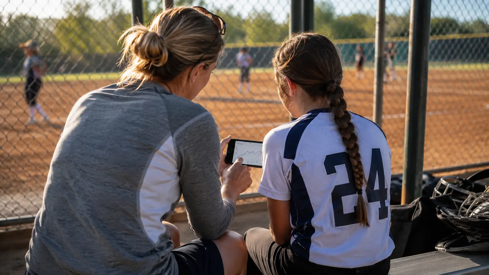 A parent and daughter sitting on a dugout bench looking at a phone screen showing a pitch velocity log