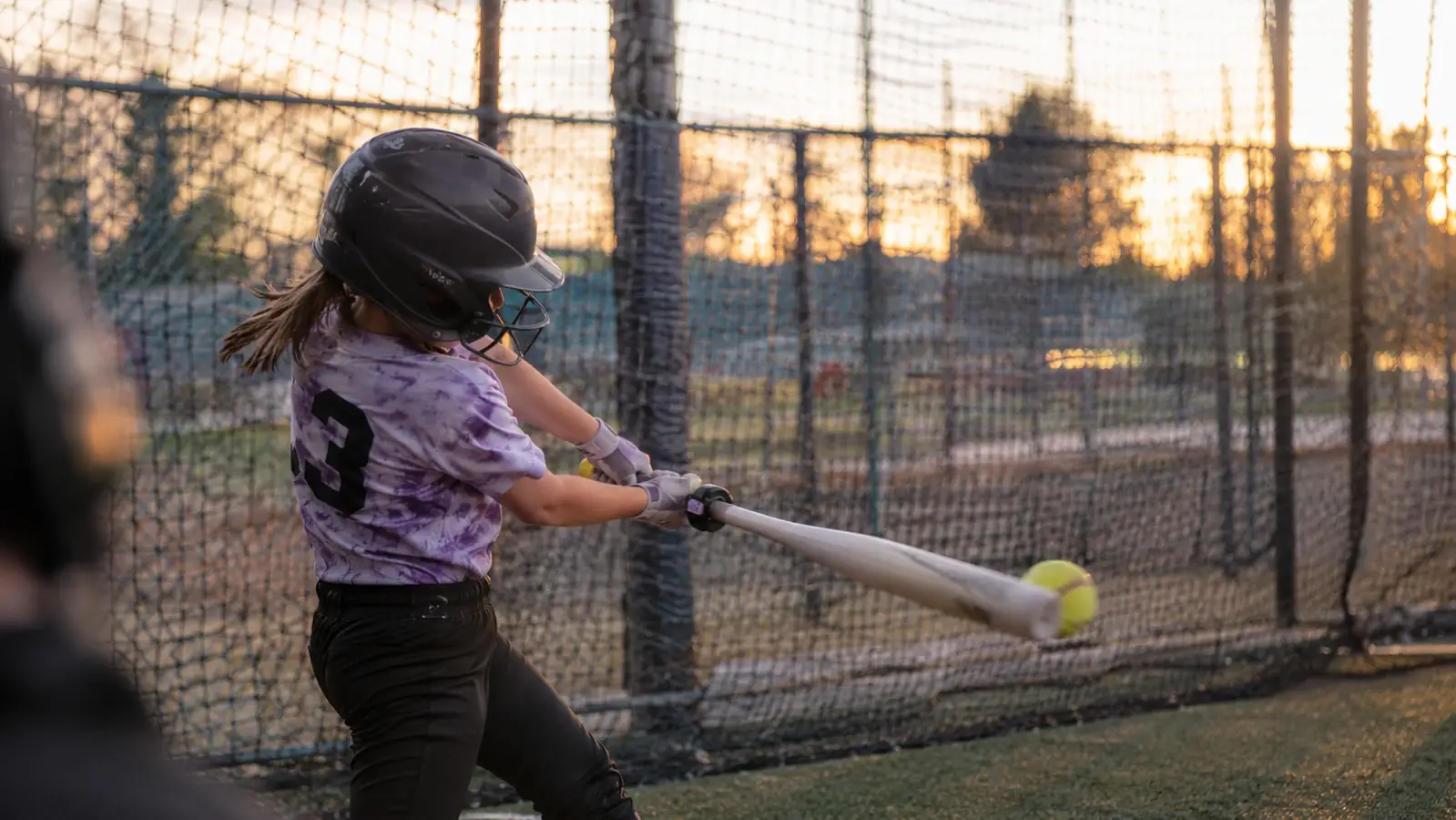 Youth softball player mid-swing in a batting cage with a small sensor visible clipped to the knob of the bat
