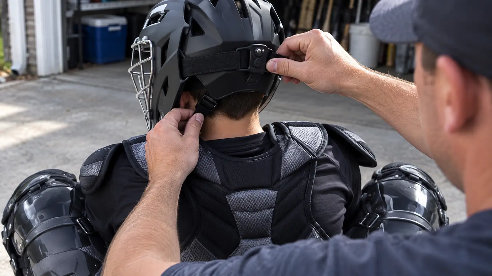 Parent's hands adjusting the chin strap and mask hinge on a catcher's helmet in a garage