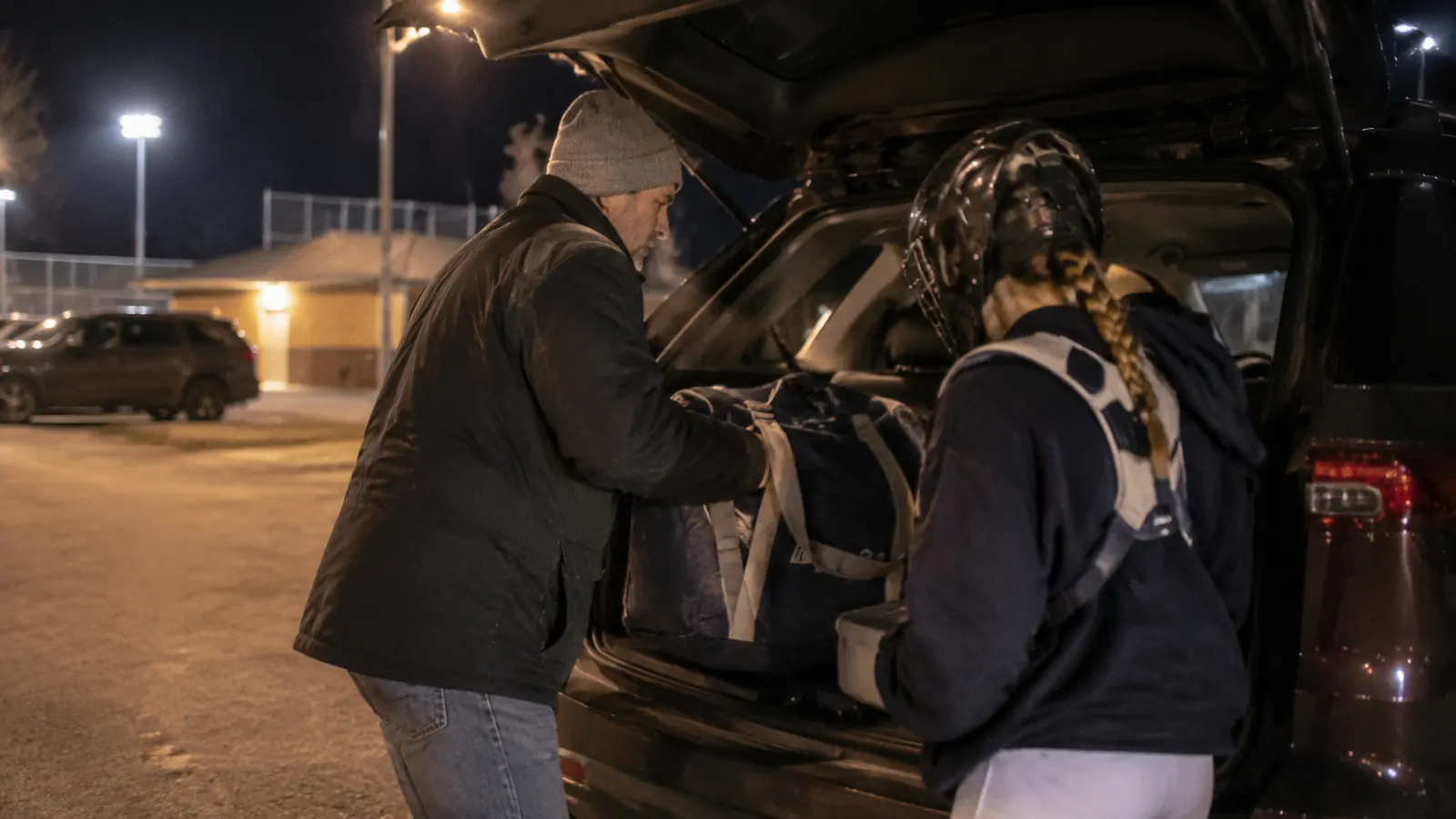 Parent and teenage daughter loading a gear bag into a car trunk in a dark, cold parking lot after practice
