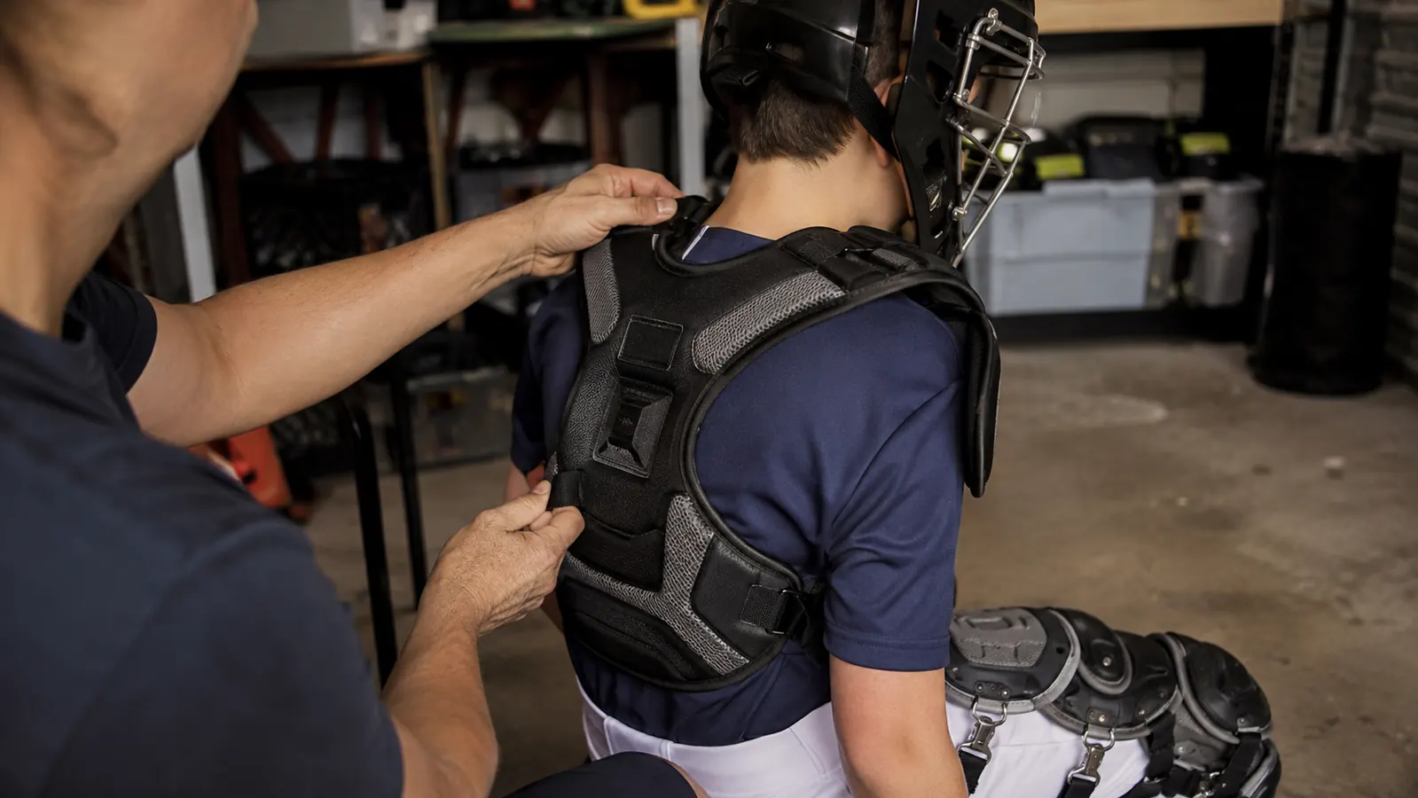 Hands buckling the chest protector harness straps over a practice jersey in a garage