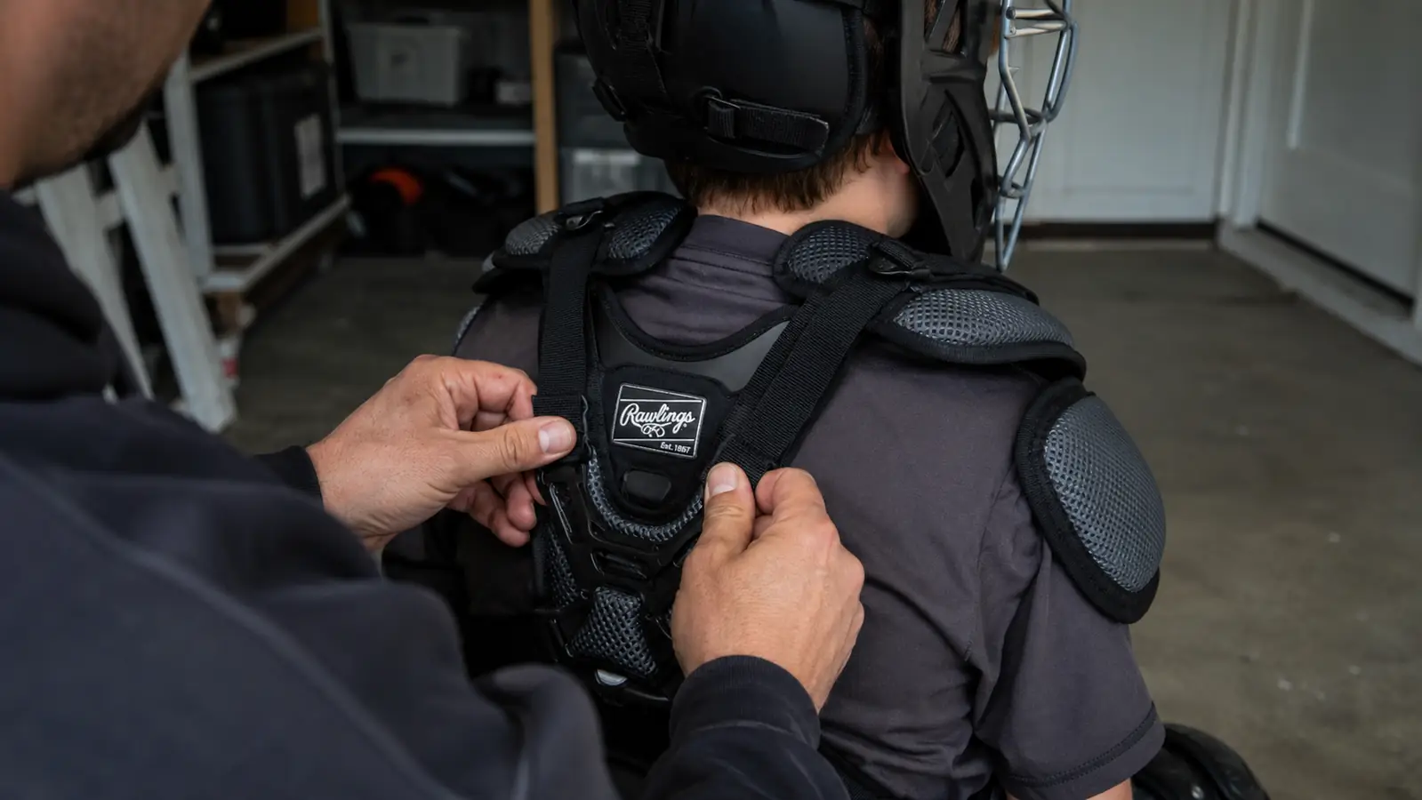 Hands buckling the chest protector harness strap on a seated youth catcher wearing the Rawlings Velo 2.0 set