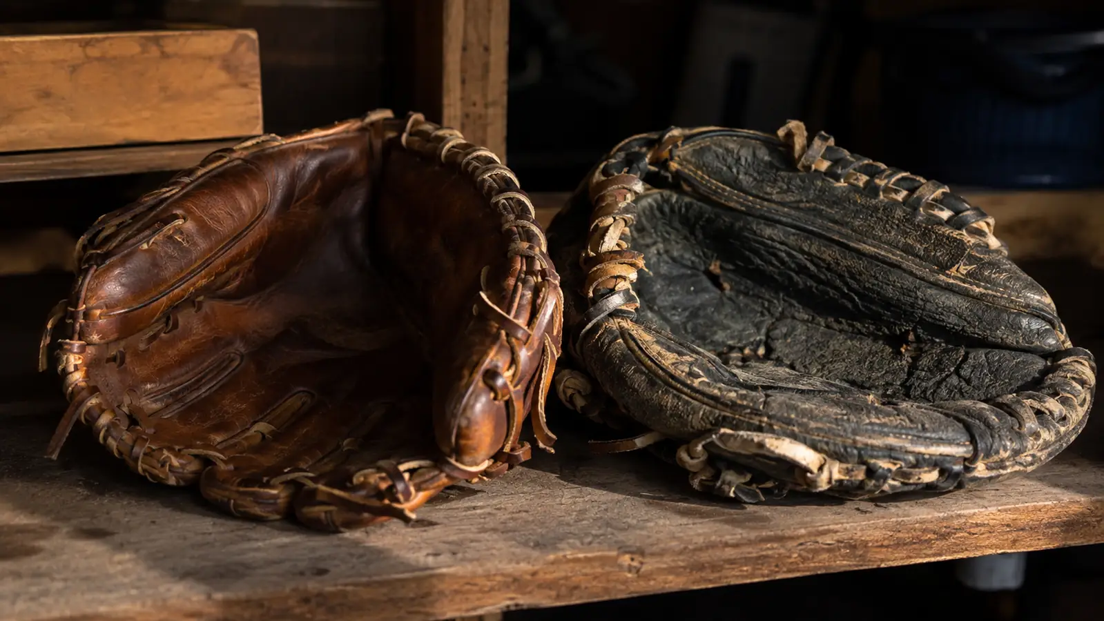A well-worn leather infield glove next to a cracked synthetic glove on a garage shelf